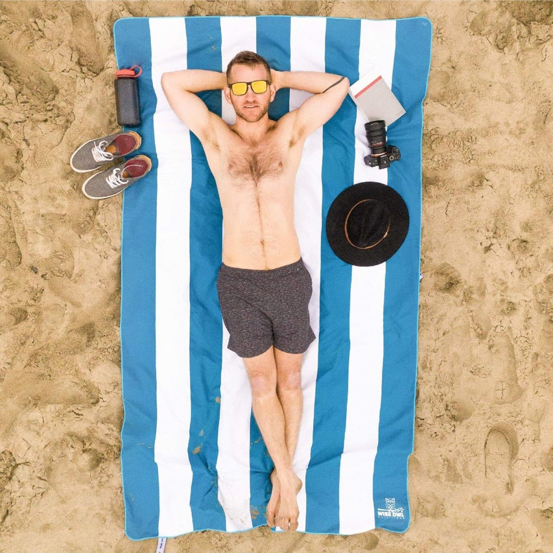 Férfi napozik kék-fehér csíkos mikroszálas strandtörölközőn a homokban, felülnézet